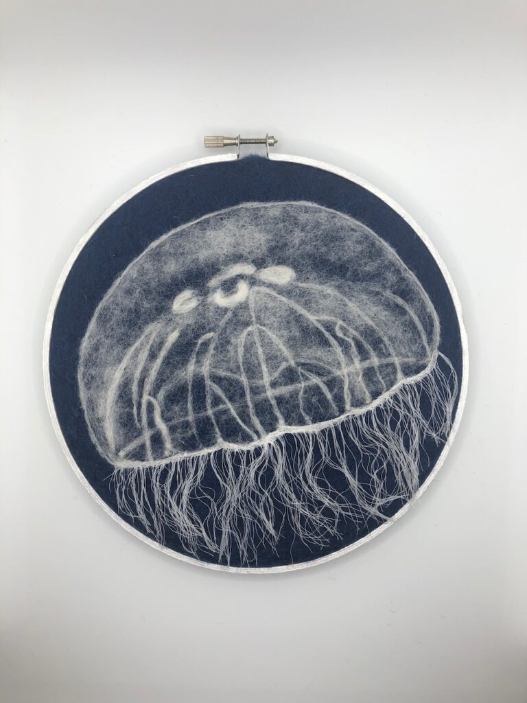 Image of a single needle felted moon jellyfish against a navy blue background. circular composition, framed in a pearlescent white embroidery hoop.